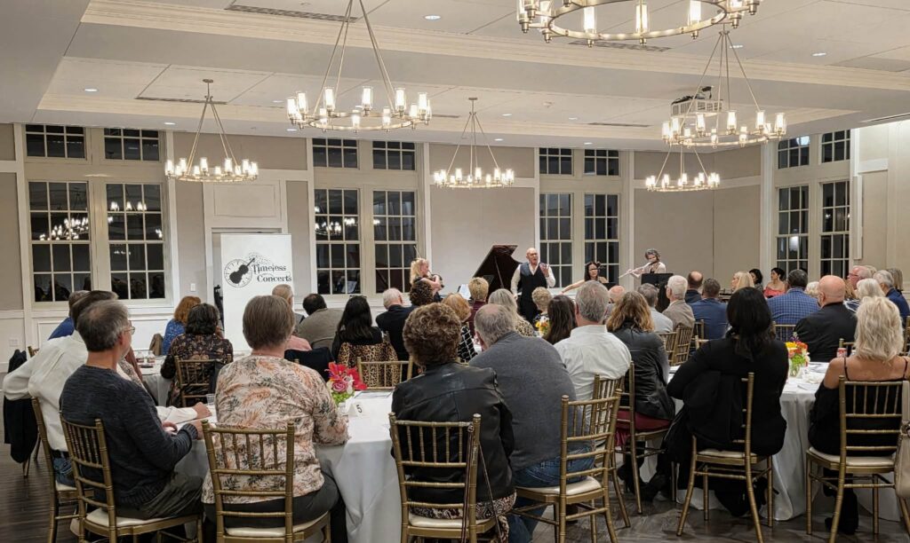 Audience seated at round tables in a banquet hall watching a small live ensemble on stage with a piano and wind players beneath large chandeliers.
