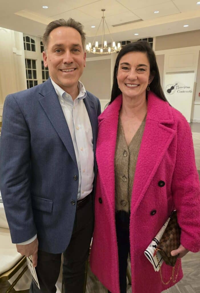 Two adults pose for a photo at an indoor event; man in a blue blazer and woman in a bright pink coat, both smiling.