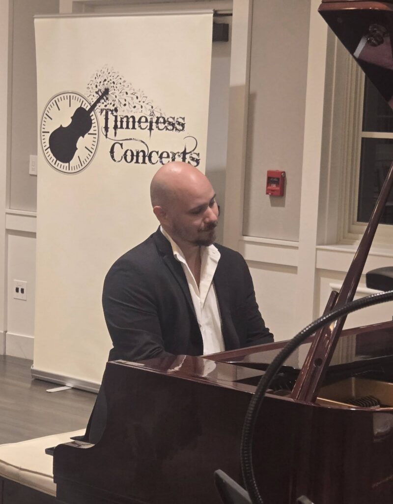 Pianist in a suit playing a grand piano at a concert indoors with a 'Timeless Concerts' banner in the background.