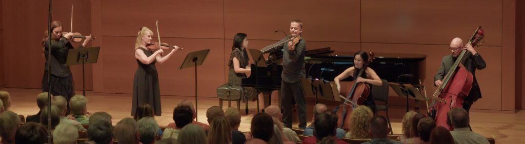 Classical string ensemble performing on stage: two violinists, pianist, cellist, and double bassist in front of an attentive audience.