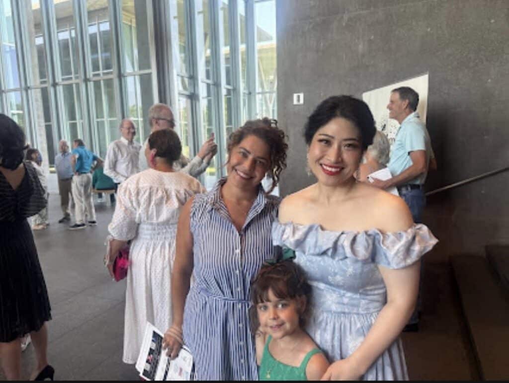 Three people pose for a photo in a glass-walled atrium: two women and a young girl in front, smiling at the camera.