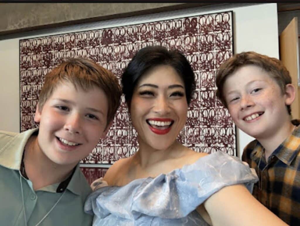 Three people pose for a cheerful selfie: a woman in a light blue off‑the‑shoulder dress between two smiling boys in casual shirts behind a patterned wall.