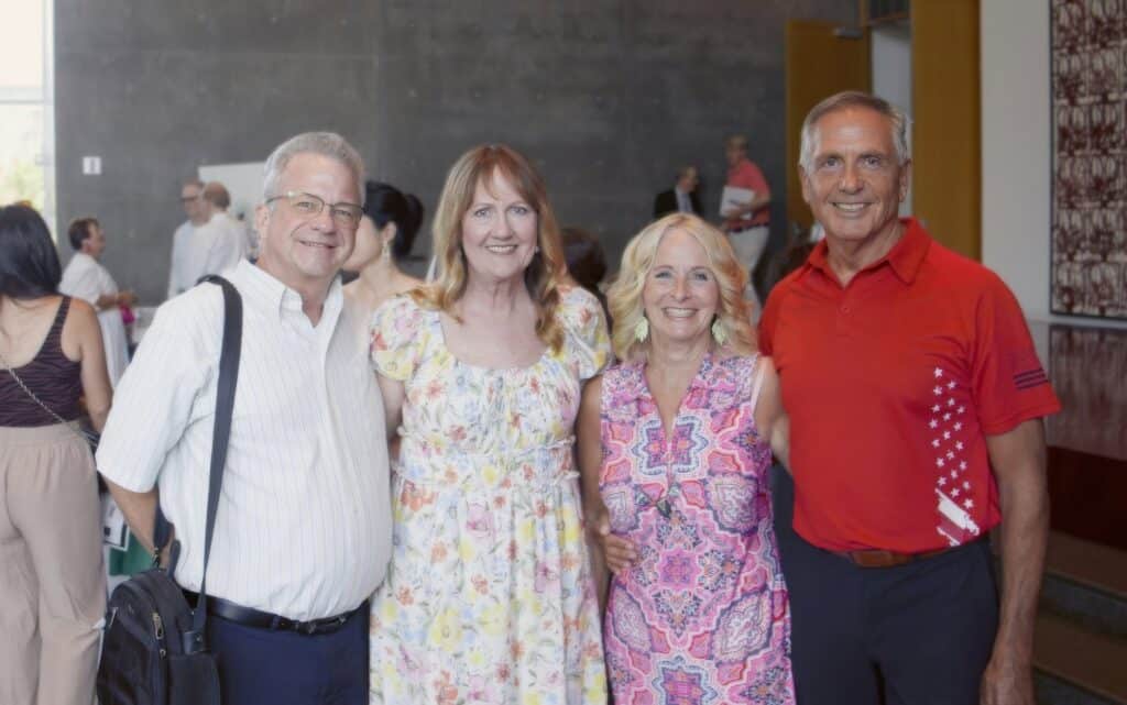 Four adults posing together for a group photo at an indoors social event; two men flank two smiling women in floral outfits, casual summer attire.