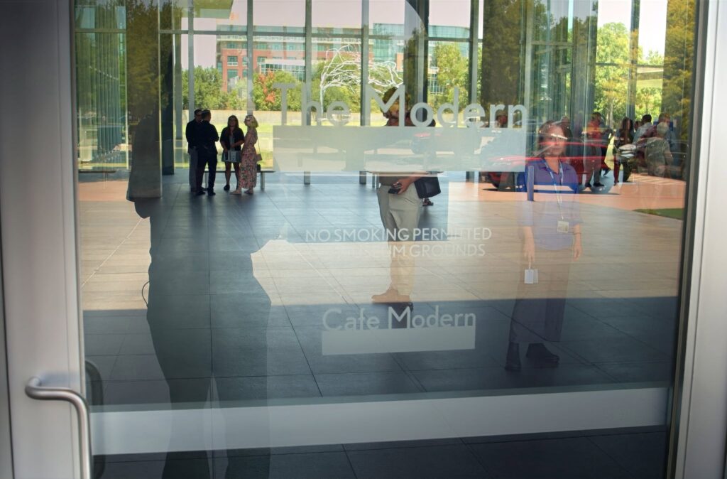 Glass entrance door with a metal handle, reflecting people outside and a modern building beyond the glass panels.