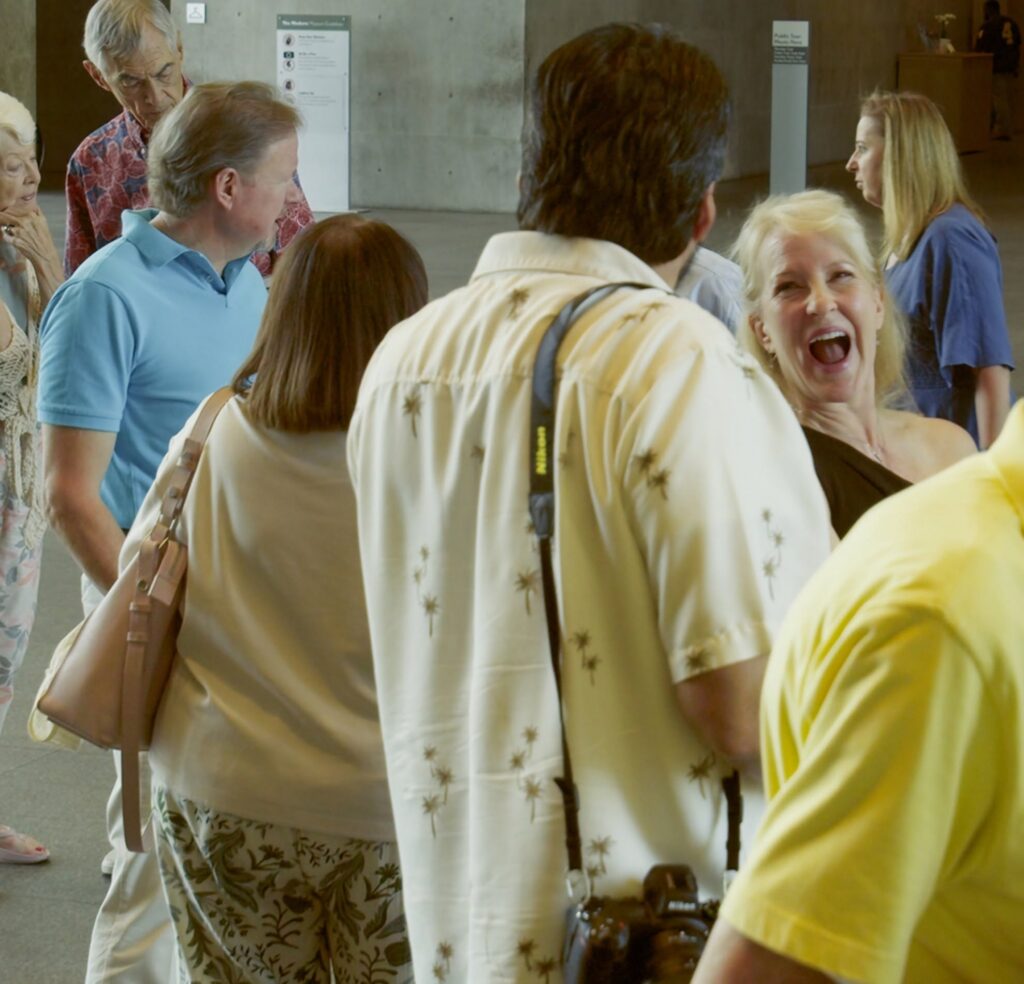 Group of adults in a casual indoor gathering; a blonde woman in the foreground laughs while others chat nearby.