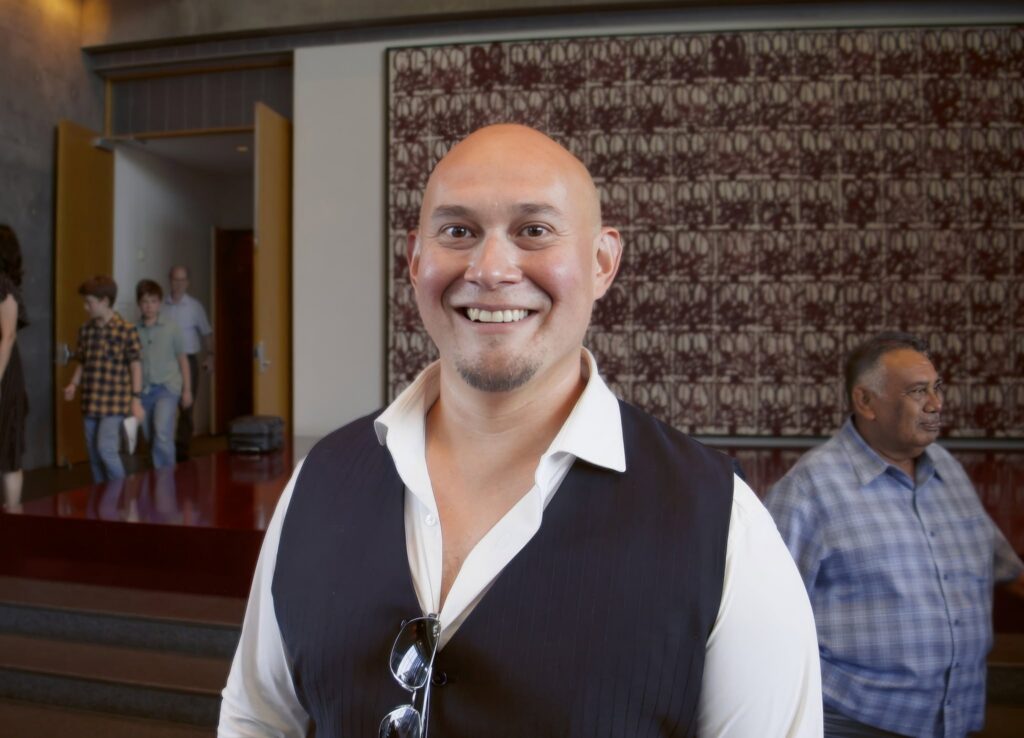 Smiling man in a white shirt and pinstripe vest facing camera at an indoor event, with patterned wall behind him and sunglasses hanging from his vest.