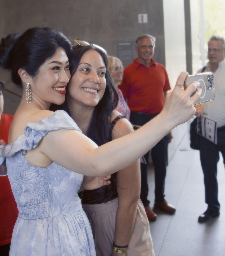 Two women in light blue dresses take a selfie, smiling, with a group of people in the background.