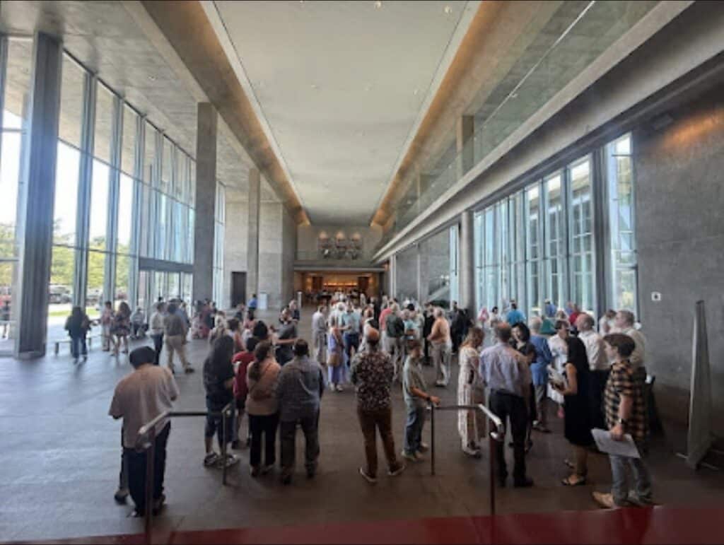 Crowd of people gathering in a bright, modern lobby with tall windows and glass railings.