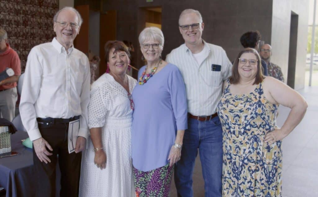 Group of five adults posing and smiling indoors at an event, dressed in casual to semi-formal clothes.