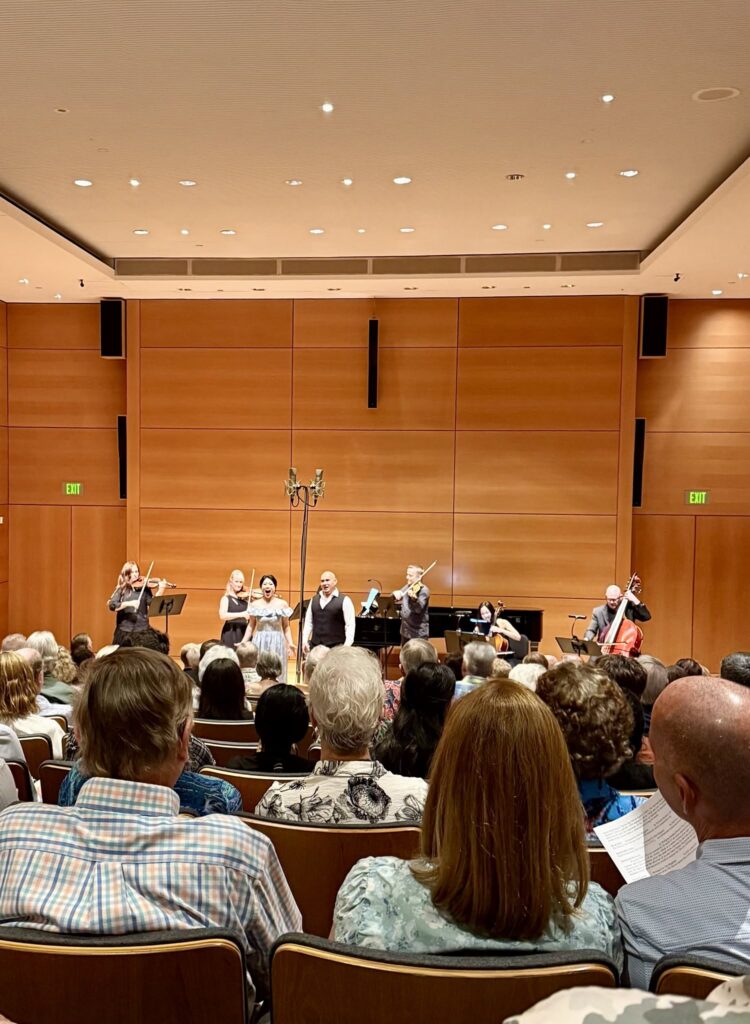 Ensemble performing on a wood-paneled concert hall stage with audience seated in the foreground.