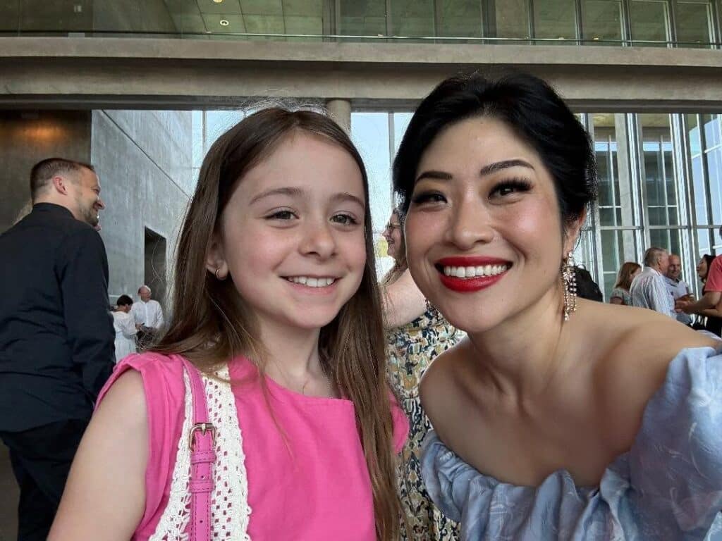 Two smiling women posing for a selfie at a social event in a modern venue, with guests in the background.