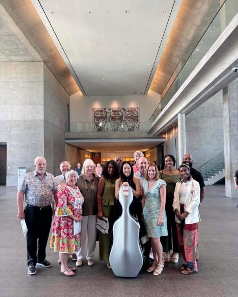 Diverse group of adults posing with a large white cello case in a modern, spacious building lobby with concrete walls and high ceilings, central to the scene.