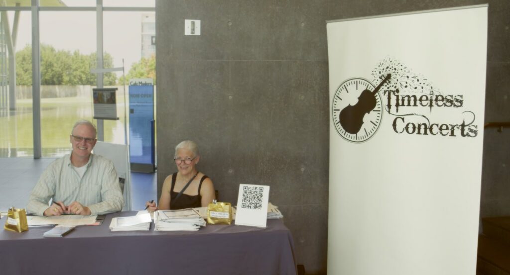 Two smiling volunteers sit at a registration desk with papers and a QR code in front, beside a large 'Timeless Concerts' banner.