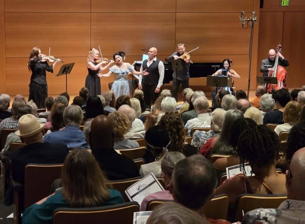 Concert hall performance featuring a female singer with an accompanying string ensemble (violinists, pianist, cellist) and an attentive audience in foreground.