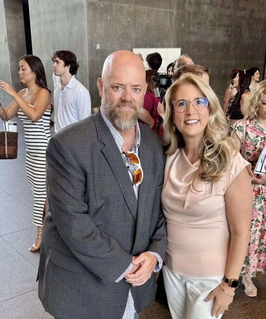 Smiling couple posing for a photo at a social event: man in a gray blazer with sunglasses hanging from his shirt, woman in a pale pink blouse, with other guests in the background.
