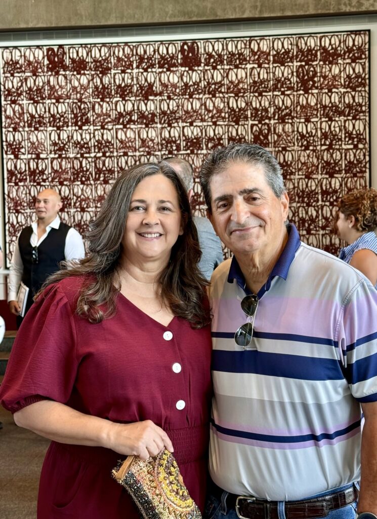Smiling couple taking a selfie indoors: woman in a burgundy dress and man in a striped polo, with a red abstract wall behind them and other guests in the background.