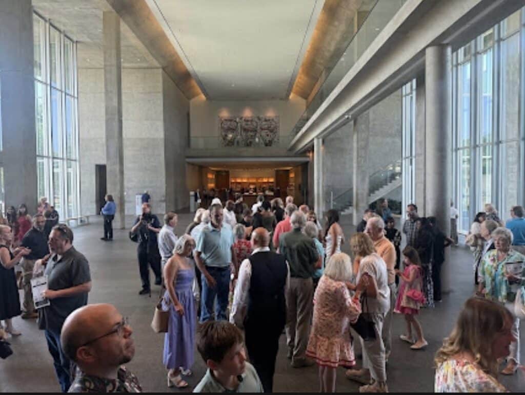 Crowd of people mingling in a bright, modern lobby with tall windows and concrete columns; reception desk visible in the background.