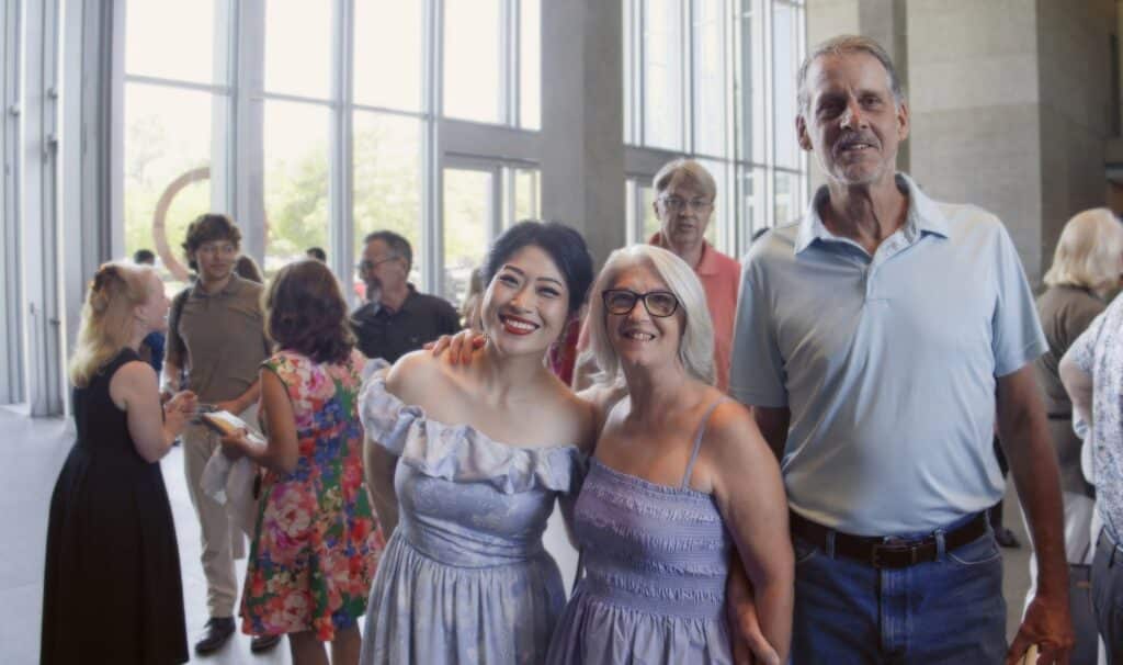 Two women in pastel dresses smile for a photo at a social event, with a tall man in a light blue shirt beside them in a bright lobby setting.