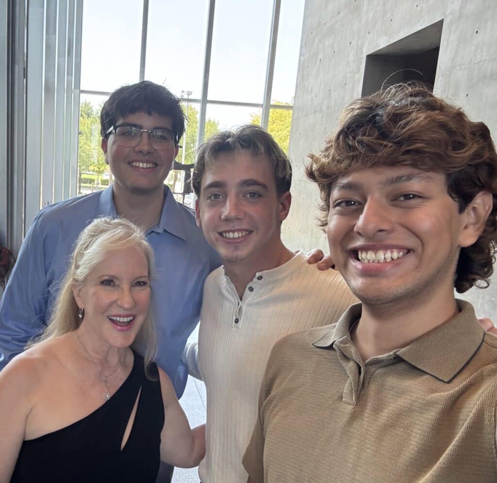 Group of four people taking a selfie indoors by tall windows, smiling at the camera in a bright lobby.