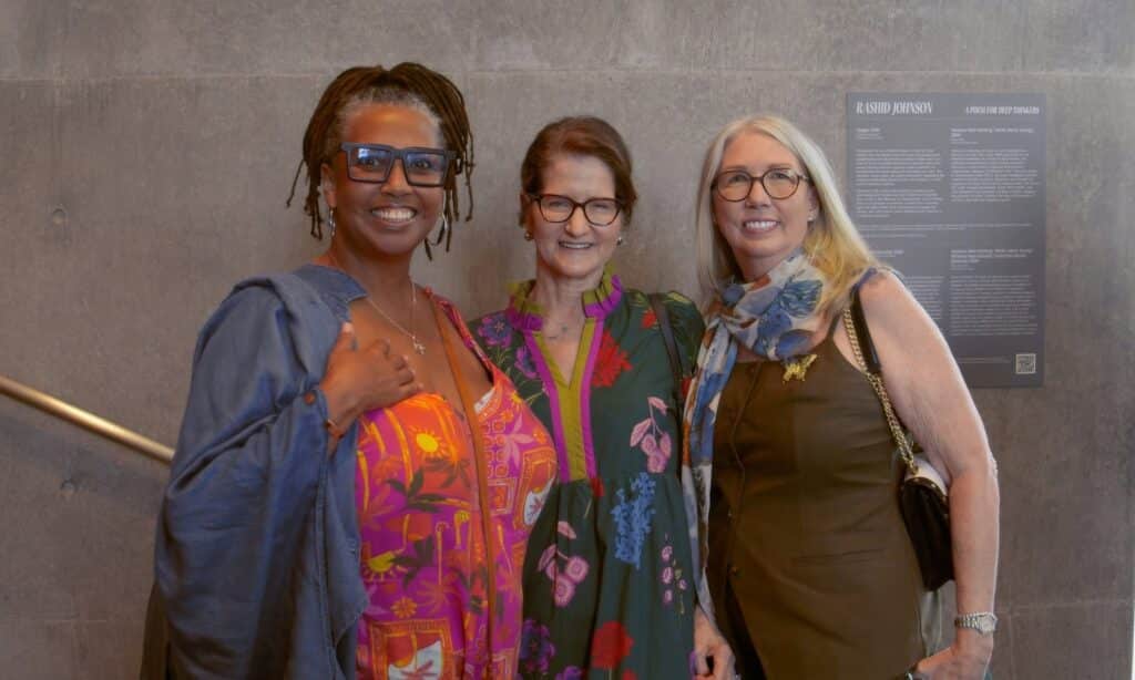Three women smiling at the camera, standing in front of a gray wall with a poster nearby.