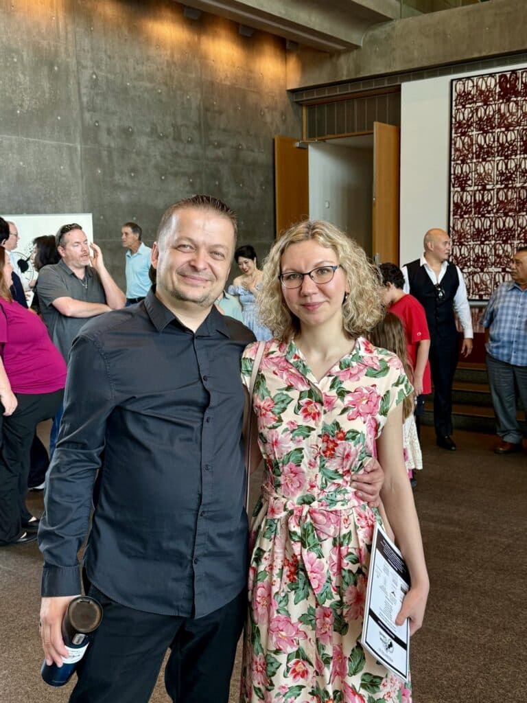 Smiling man in a dark shirt and smiling woman in a floral dress pose together at a crowded indoor event.
