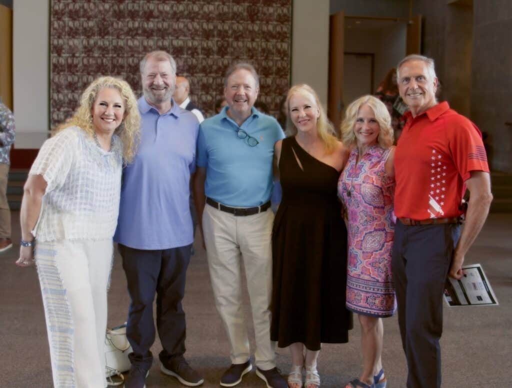 Six adults posing together indoors, smiling for the camera in a lobby setting behind them a patterned wall is visible.