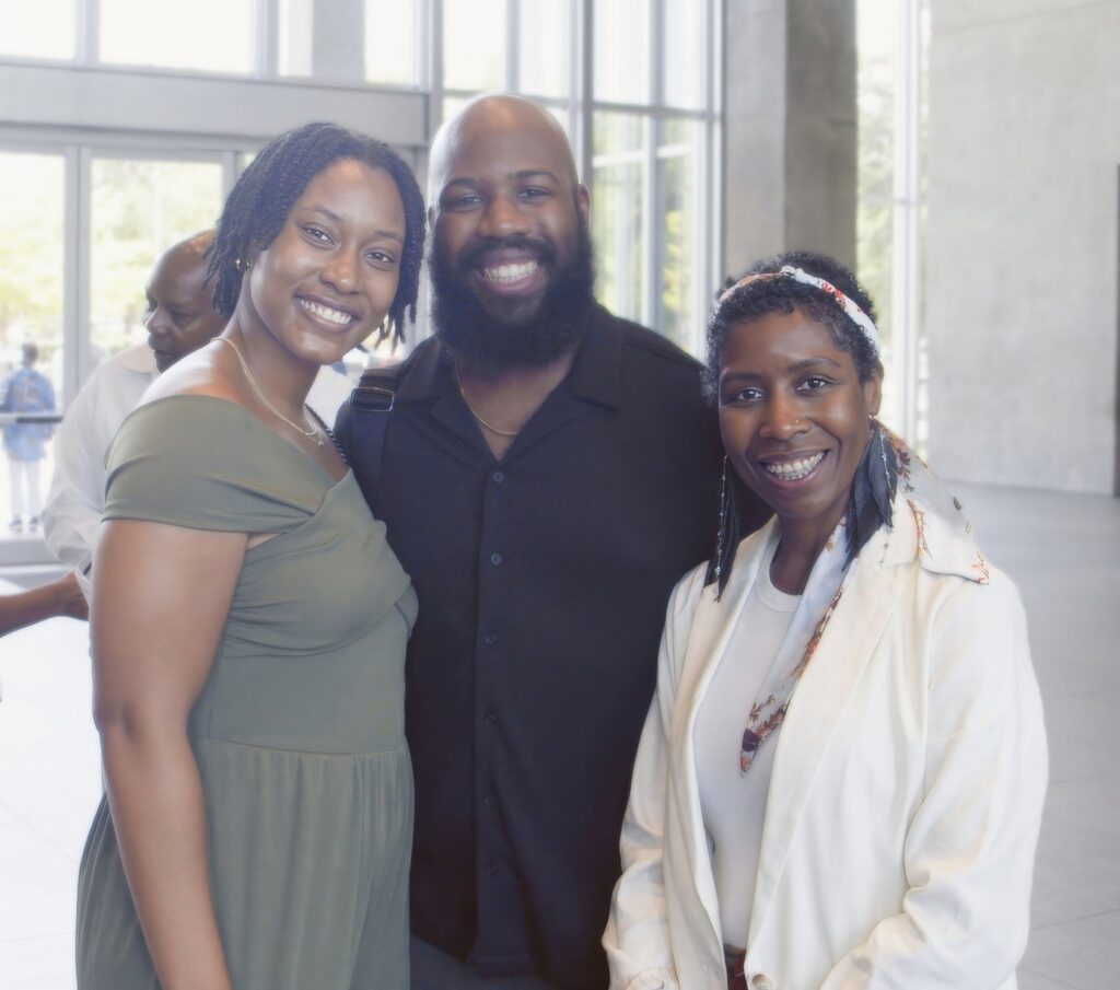 Three friends posing for a selfie in a bright lobby, smiling at the camera with arms around each other.