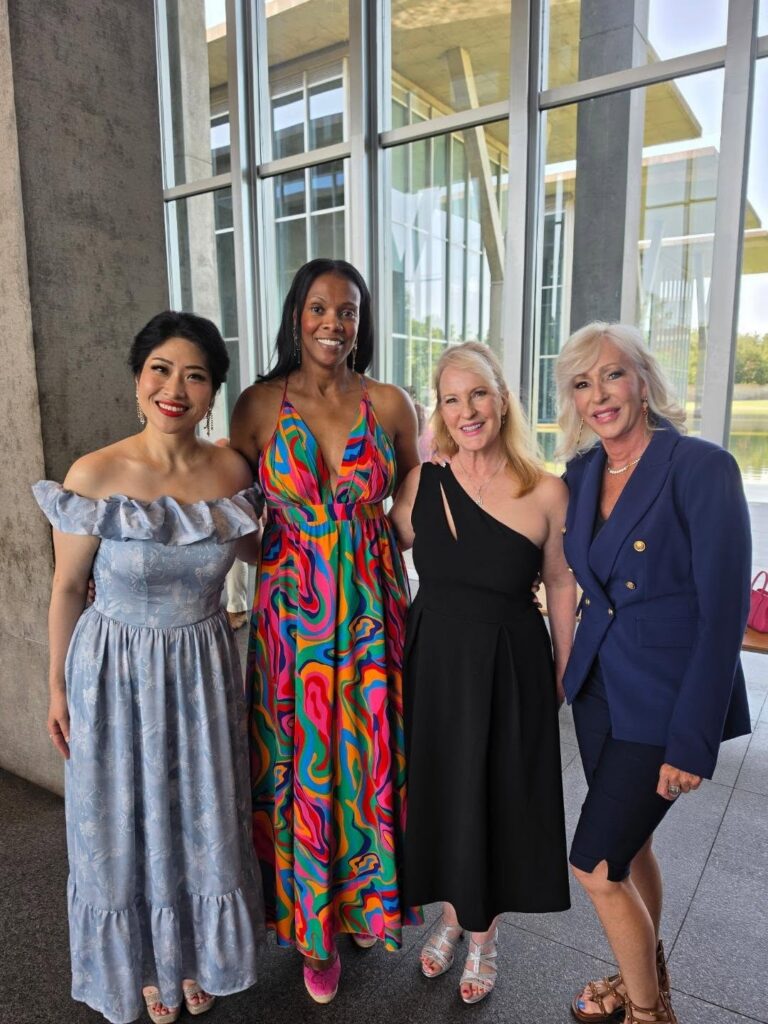 Four women pose together at an event in a bright, glass-walled venue, dressed in coordinated formal outfits.